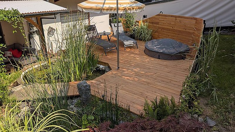 Holzterrasse mit Liegen, Sonnenschirm und Pflanzen, umgeben von einem kleinen Teich und Garten.