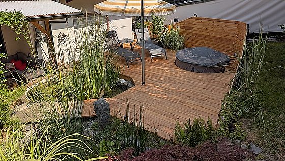 Holzterrasse mit Liegen, Sonnenschirm und Pflanzen, umgeben von einem kleinen Teich und Garten.