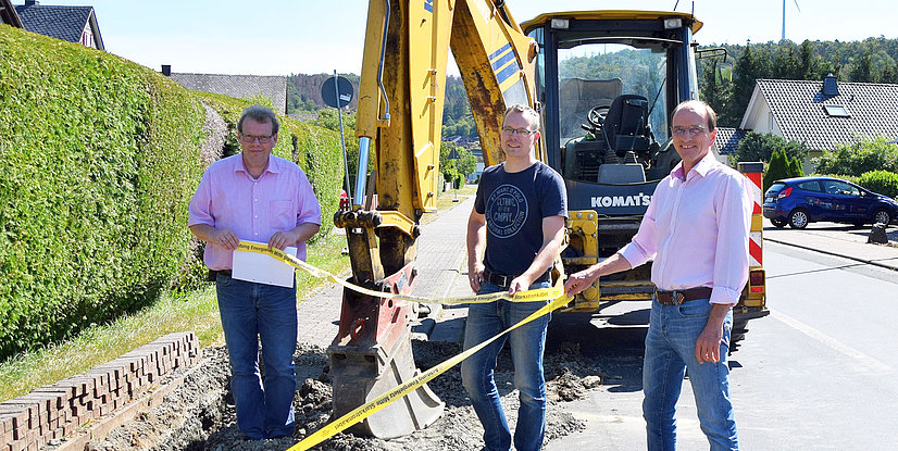 Drei Männer stehen an einer Baustelle mit einem Bagger und halten ein Sicherheitsband.