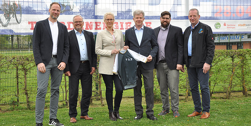 Gruppenszene mit sechs Personen, die ein TDE-Trikot halten, vor einem Zaun und einem Banner im Hintergrund.