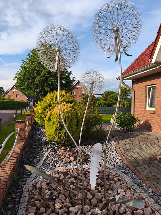 Skulptur von drei stilisierten Pusteblumen aus Metall im Garten, umgeben von Steinen und Pflanzen.