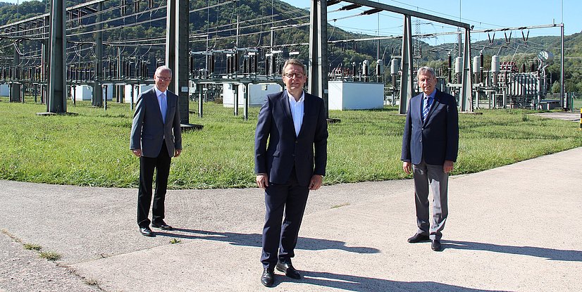 Drei Männer in formeller Kleidung stehen vor einer elektrischen Umspannstation in einem grünen Landschaftsbereich.