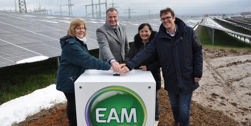 Vier Personen stehen vor Solarmodulen und betätigen gemeinsam einen Knopf auf einem weißen Kasten mit grüner EAM-Logo.
