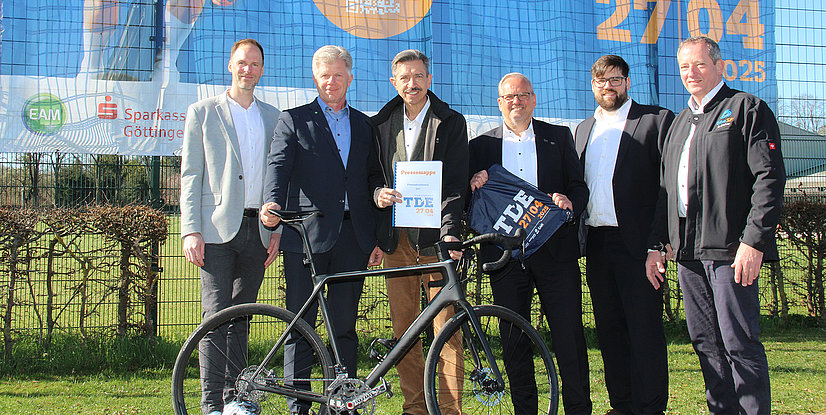 Sechs Personen stehen vor einem großen Banner, vor ihnen steht ein modernes Rennrad und sie halten eine TDE-Urkunde.