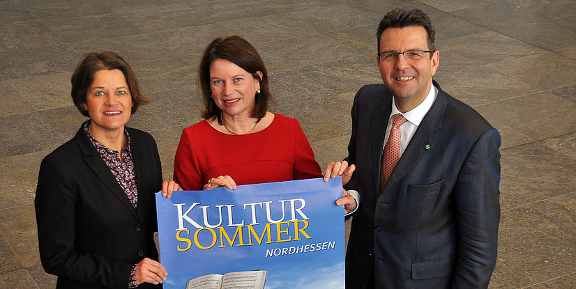 Drei Personen stehen in einem Raum und halten ein Plakat mit der Aufschrift KULTURSOMMER NORDHESSEN