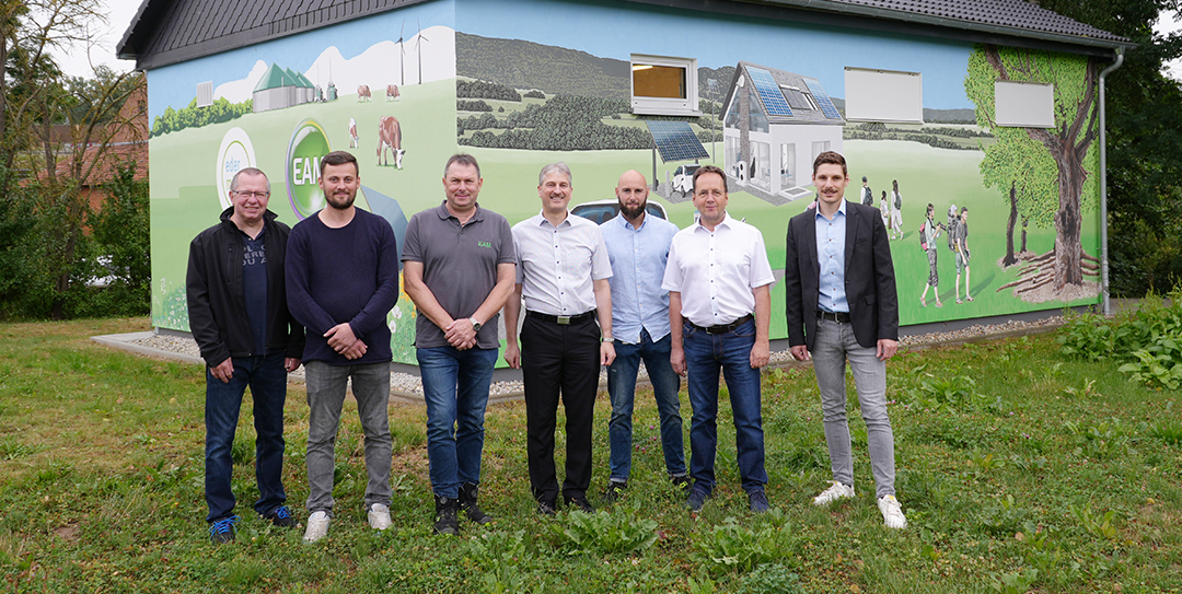 Eine Gruppe von sieben Männern (Namen und Funktion siehe Bildunterschrift) steht vor der neuen bemalten Schaltstation. Die Motive reichen von einem Haus mit PV-Anlage über ein E-Auto, Windkraft- und Biogasanlagen bis hin zu Menschengruppen, Bäumen und Kühen in der Natur.