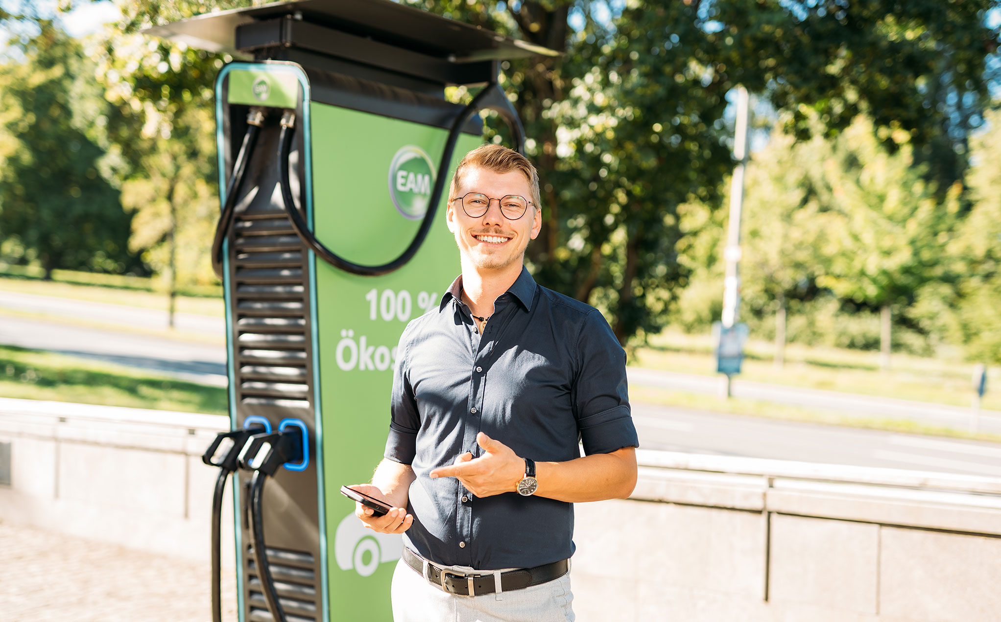 Lächelnder Mann mit Smartphone neben einer grünen Ladestation für Elektroautos im Freien.