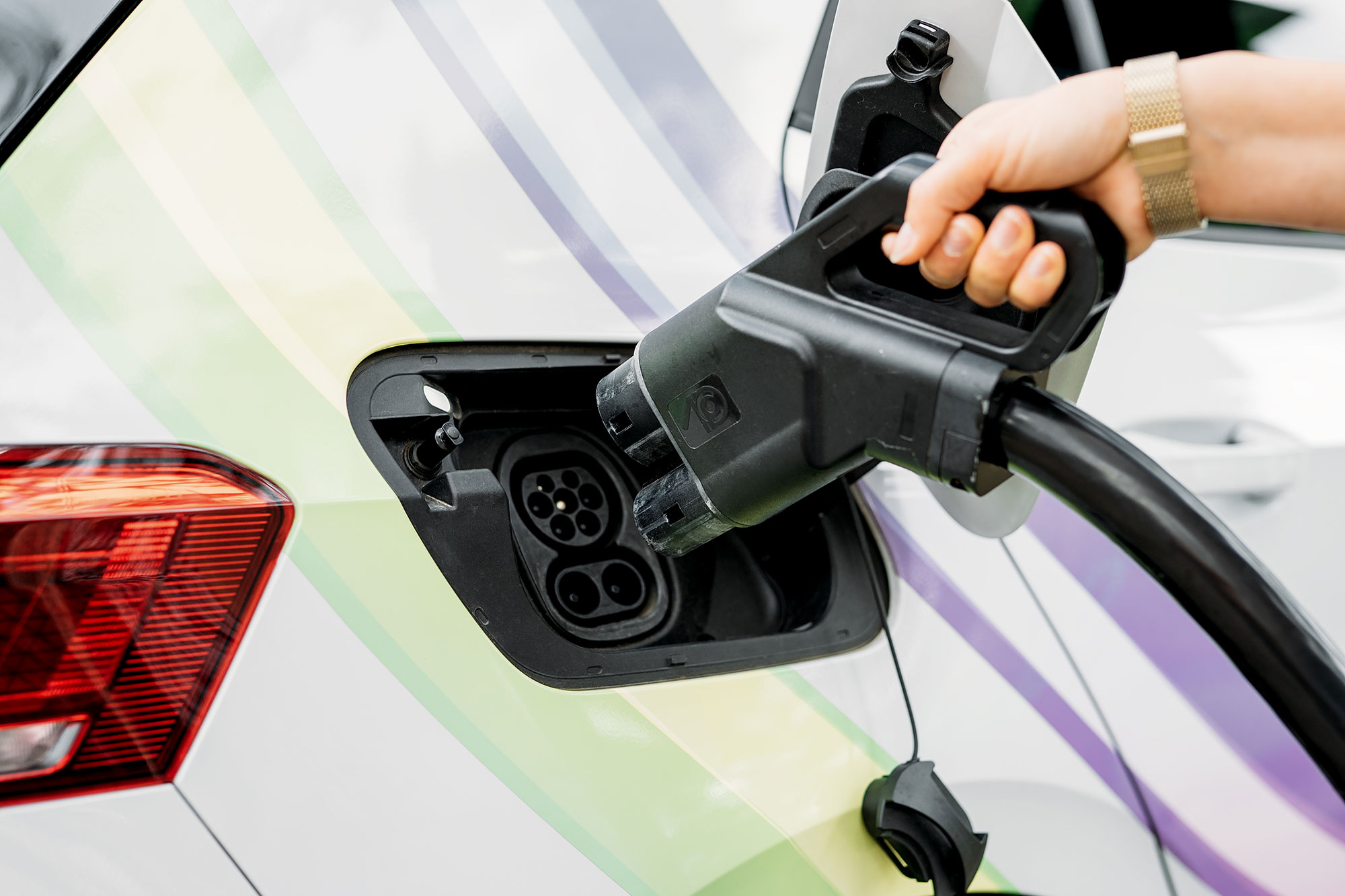 Hand, die einen Ladestecker in die Ladebuchse eines Elektroautos einführt. Sichtbare Autooberfläche mit buntem Design.