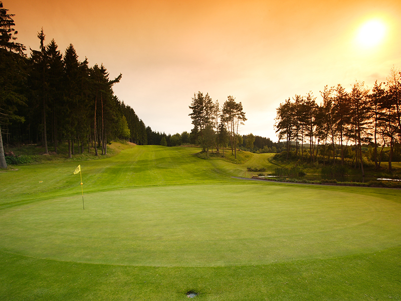 Grünes Golfplatz-Setting mit Fahne, umgeben von Bäumen und sanften Hügeln unter einem orangefarbenen Himmel.