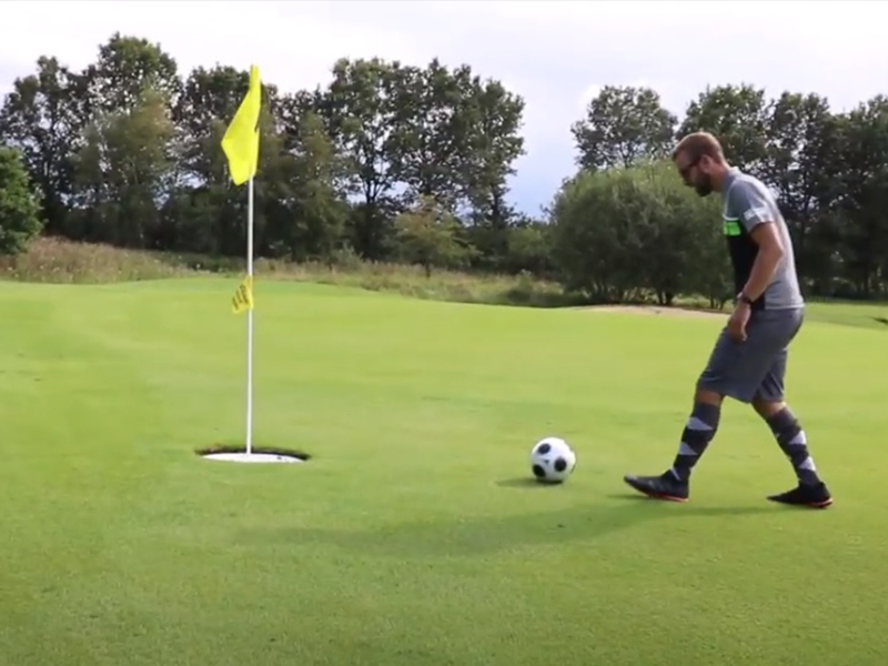 Ein Mann in Sportkleidung zieht auf einem grünen Platz einen Fußball in Richtung einer Fahne am Loch.