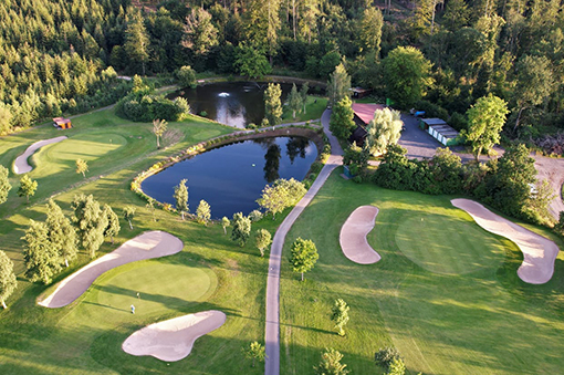 Luftaufnahme eines Golfplatzes mit Grünflächen, Sandbunkern, einem Teich und umliegenden Bäumen.