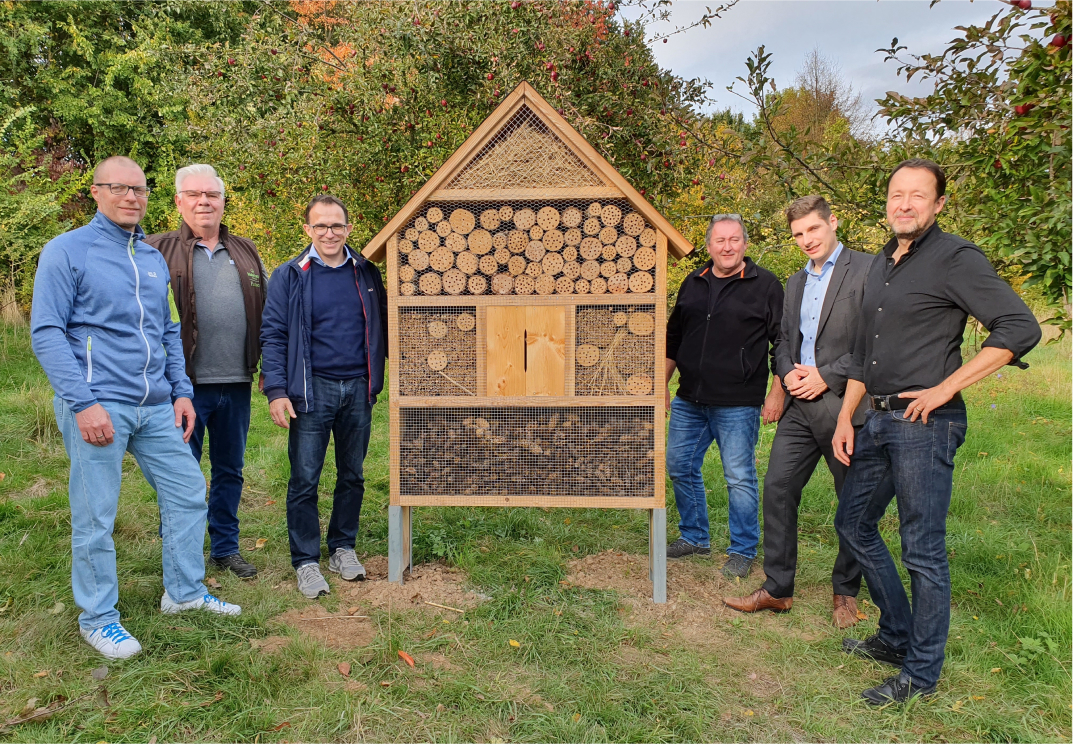 Sechs Personen stehen neben einem Insektenhotel in einer herbstlichen Landschaft.