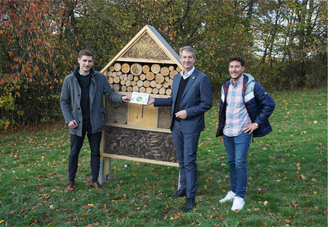 Drei Personen stehen vor einem Insektenhotel in einem herbstlichen Garten und halten ein Schild.