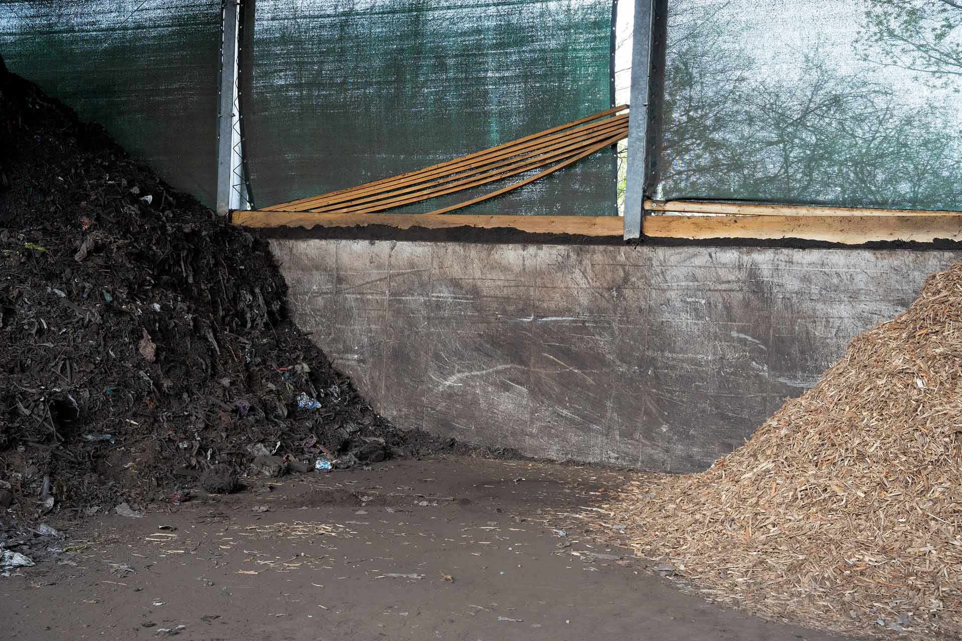 Auf einem Kompostgelände sind zwei große Hügel aus organischen Abfällen und Holzschnitzeln unter einem grünen Netz zu sehen.