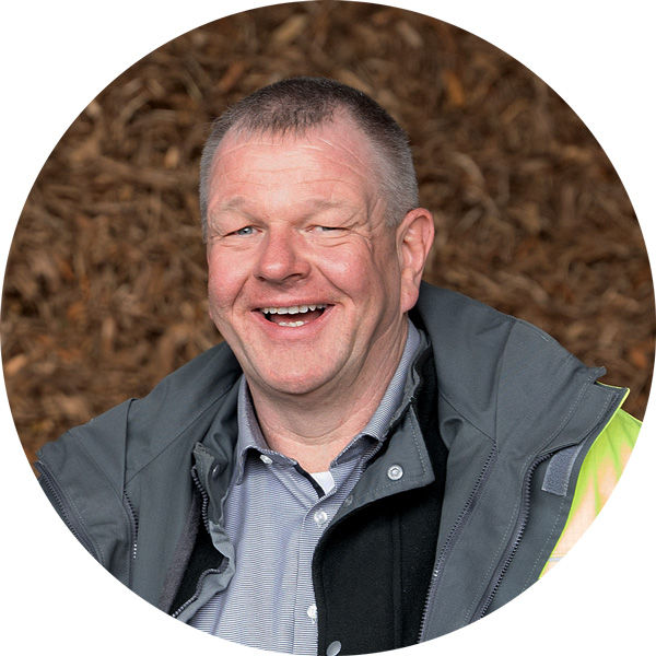 Lächelnder Mann in Arbeitskleidung mit grauer Jacke vor einem Hintergrund aus Holzspänen.