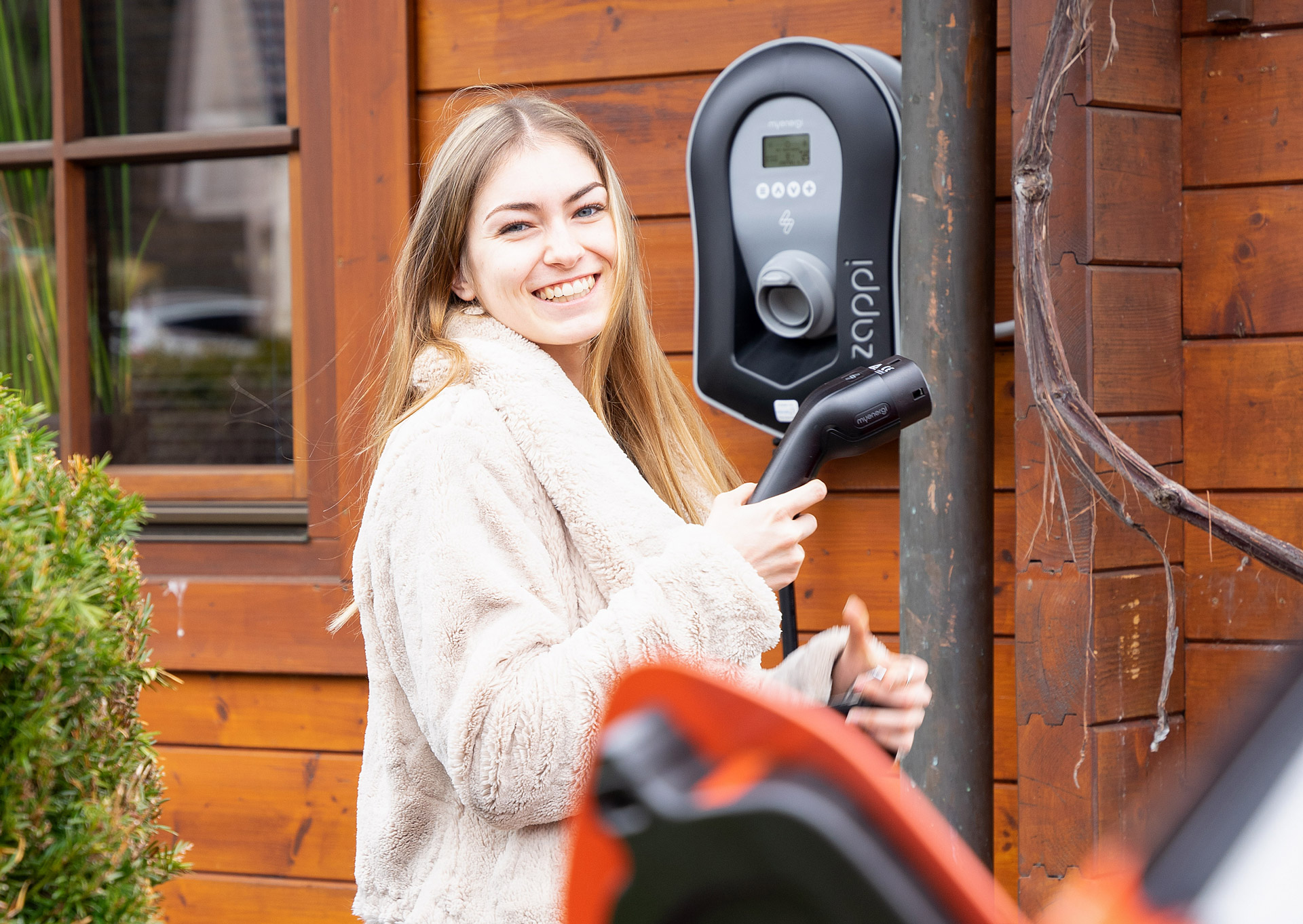 EAM Klima WWOI Tochter Seidl Junge Frau lächelt, während sie einen Ladegriff an einer Ladestation für Elektroautos hält, vor einem Holzhaus.