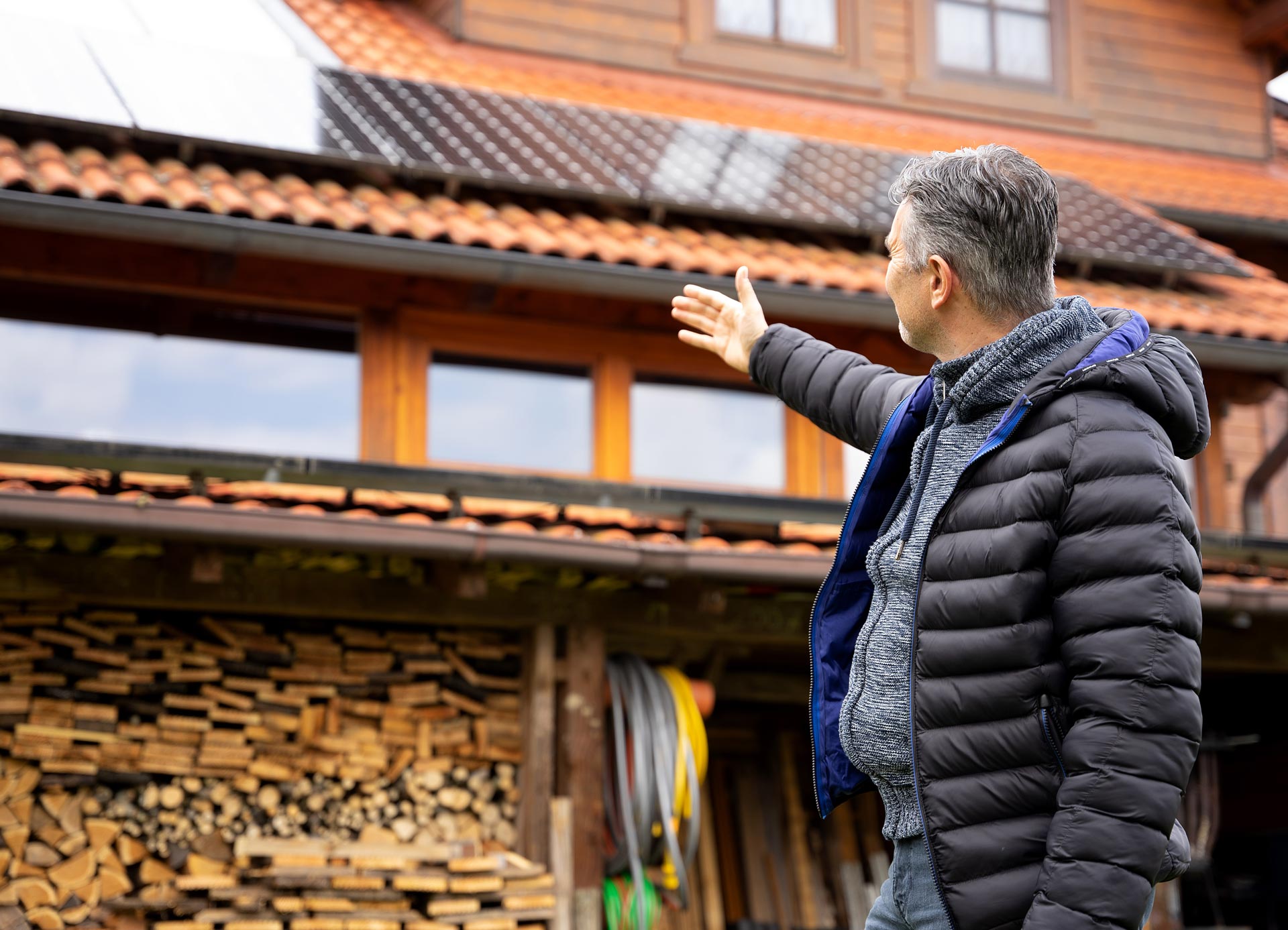 Mann steht vor einem Holzhaus und zeigt auf die Solarpanels auf dem Dach. Holzstapel und Werkzeuge im Hintergrund sichtbar.