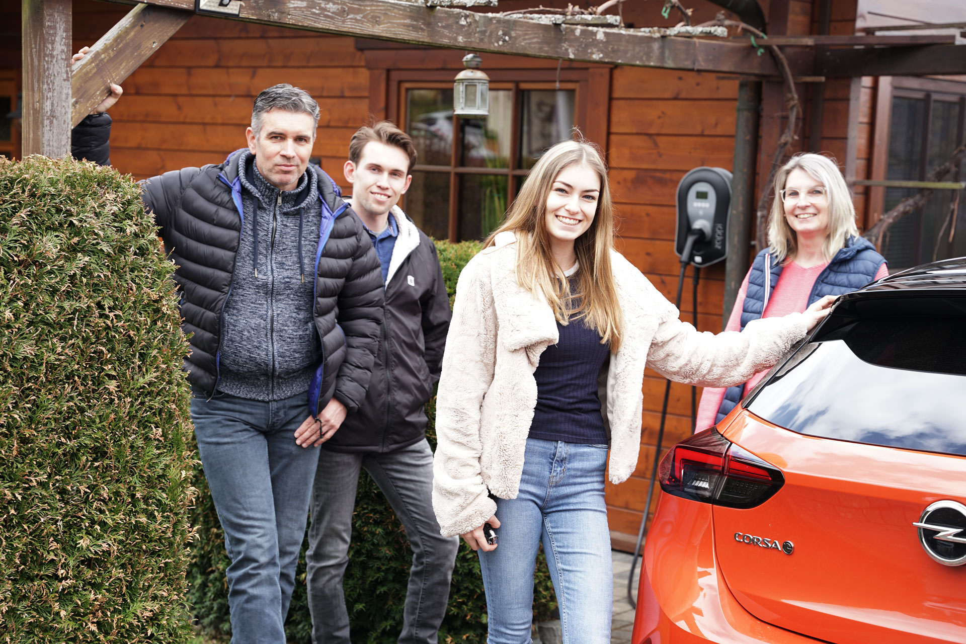 Vier Personen stehen lächelnd vor einem orangefarbenen Auto vor einem Holzhaus, umgeben von grünen Pflanzen.