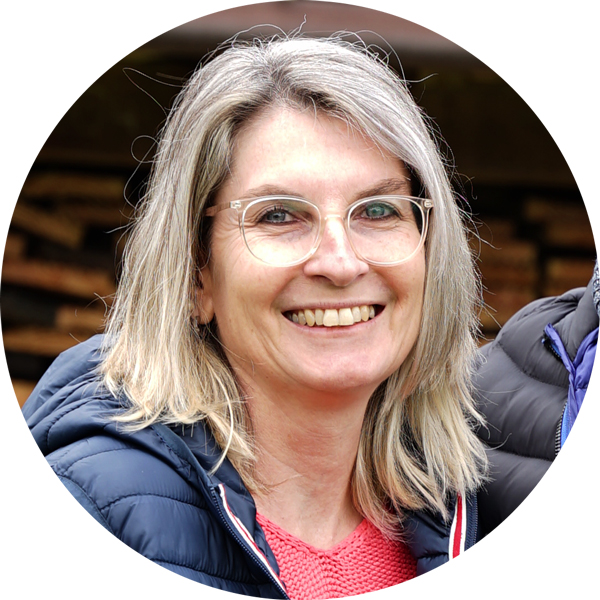 Eine lächelnde Frau mit blonden Haaren und Brille, trägt eine Jacke, vor unscharfem Hintergrund.