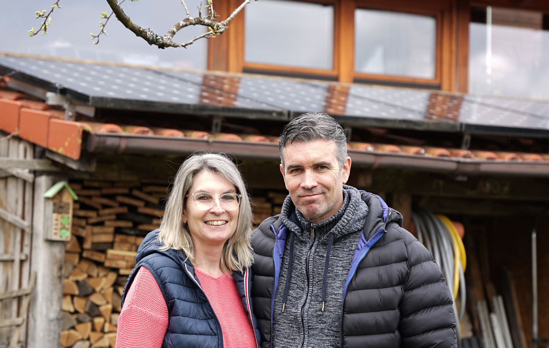 Paar steht lächelnd vor einem Holzstapel, im Hintergrund ein Haus mit Solarpanelen und grünen Pflanzen.