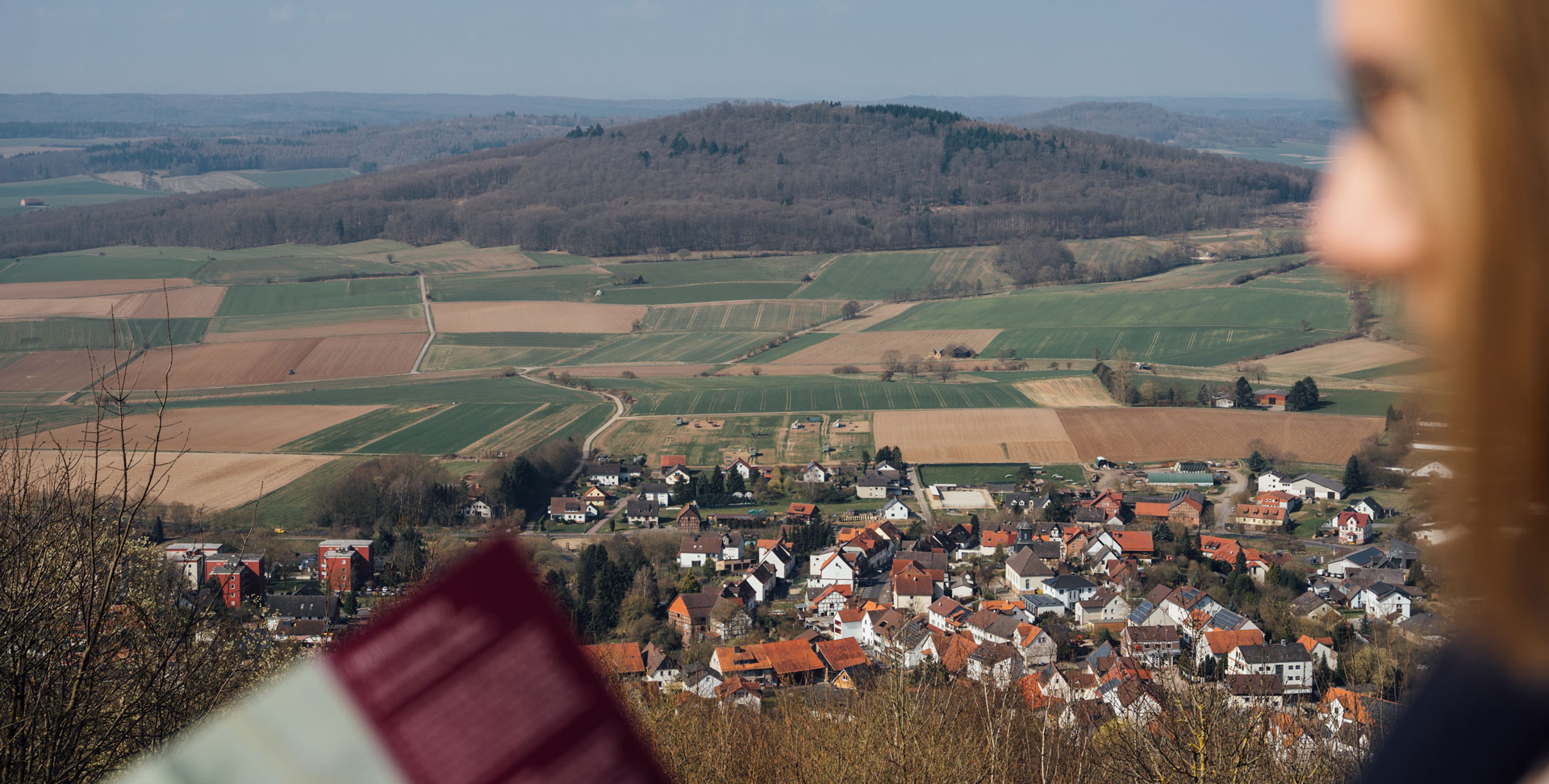 Blick auf eine ländliche Landschaft mit Feldern und einem Dorf im Vordergrund, während eine Person am Rand steht.