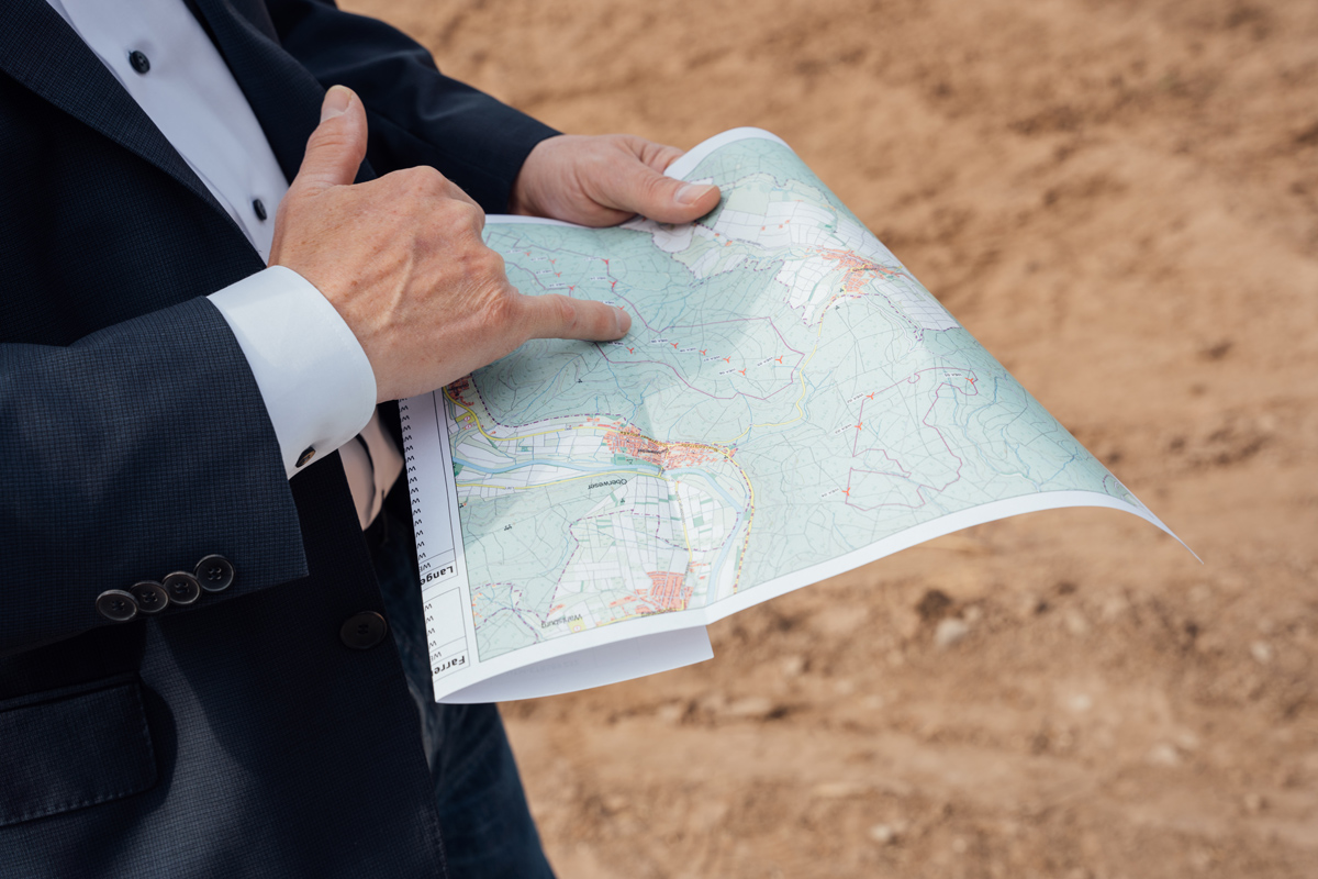Person in Anzug hält eine Landkarte und zeigt mit dem Finger auf einen bestimmten Punkt. Hintergrund ist brauner Boden.
