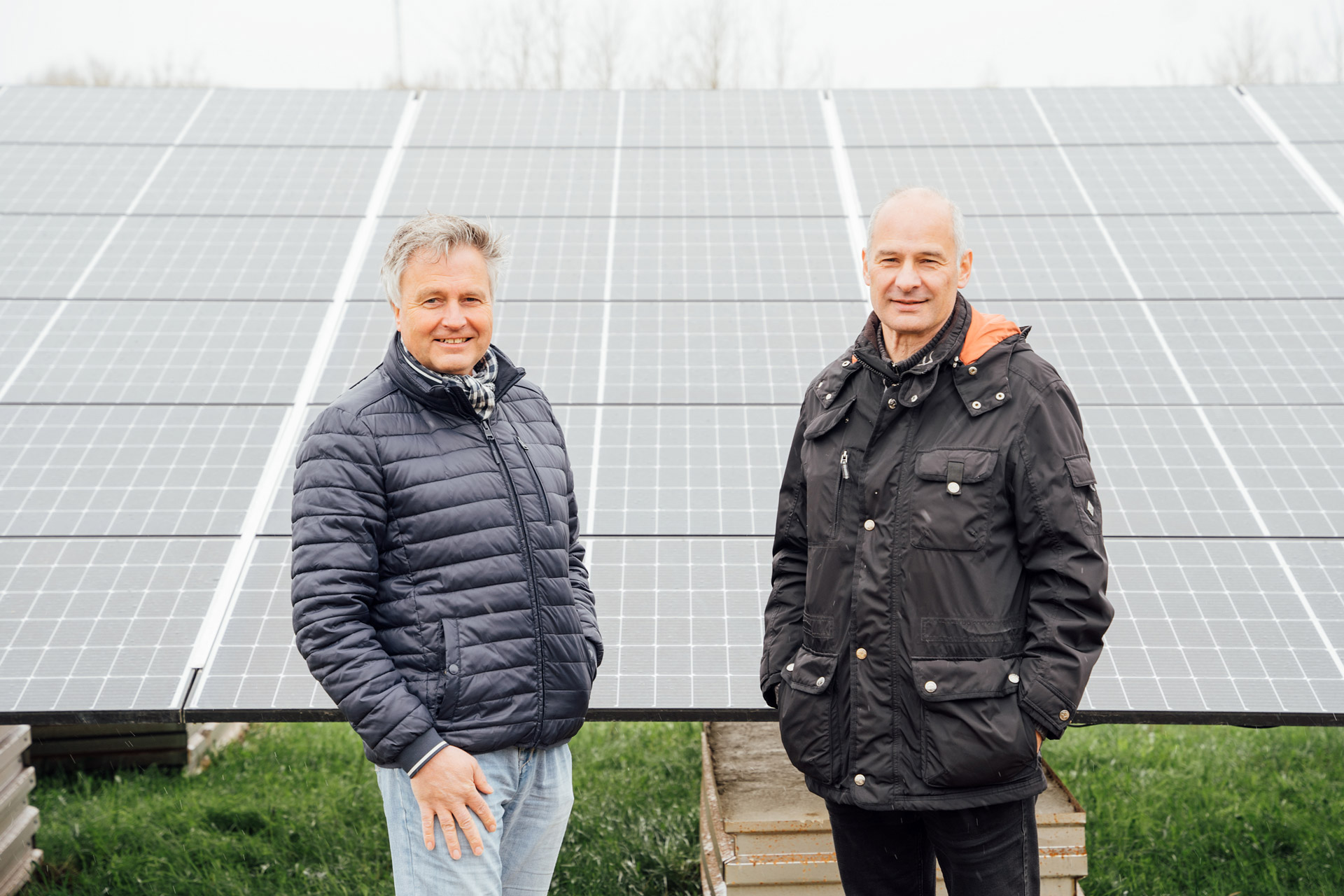 Zwei Personen stehen vor Solarpaneelen auf einem grünen Feld, über dem ein grauer Himmel zu sehen ist.