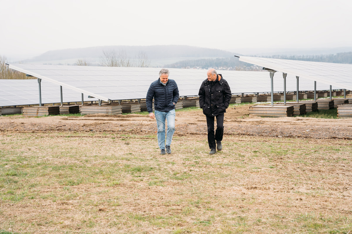 Zwei Personen gehen an Solarpanelen vorbei, umgeben von einer grünen Wiese unter einem grauen Himmel.
