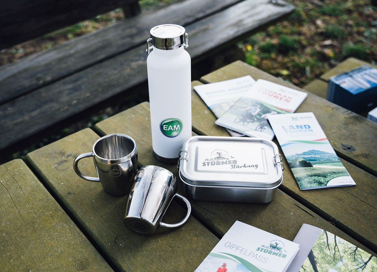 Stainless steel cups, a water bottle, a lunchbox, and brochures arranged on a wooden table outdoors.