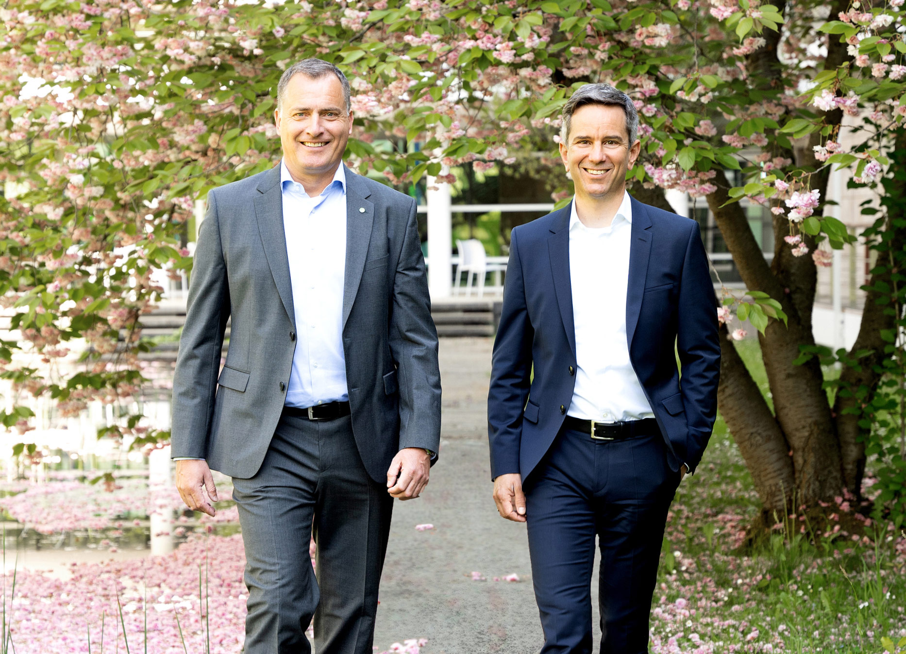 Zwei Männer in Anzügen gehen lächelnd einen Blumengartenweg entlang, umgeben von blühenden Bäumen.