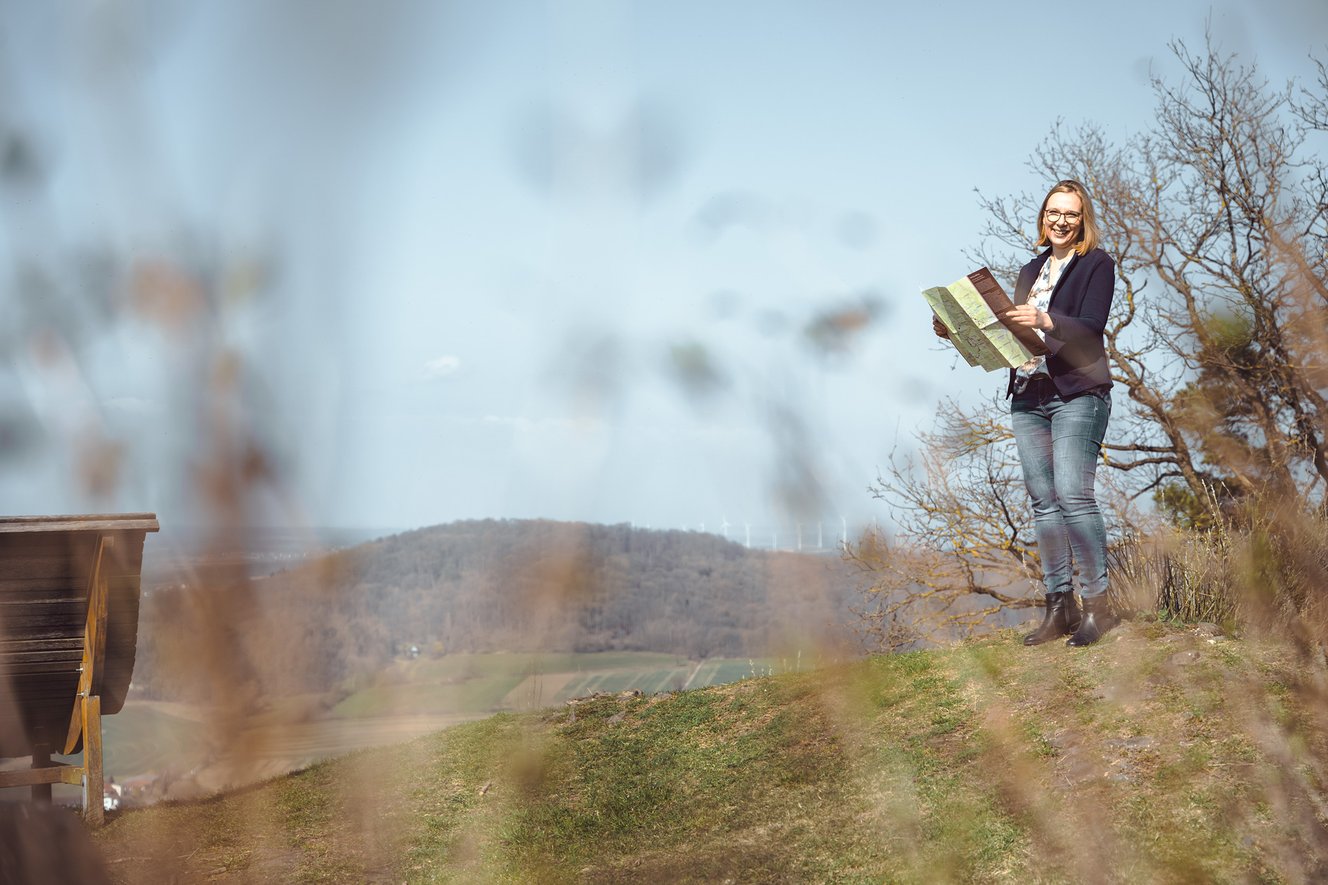 Frau mit Karte steht auf einem Hügel, umgeben von Gras und Bäumen, mit weitem Blick auf die Landschaft.