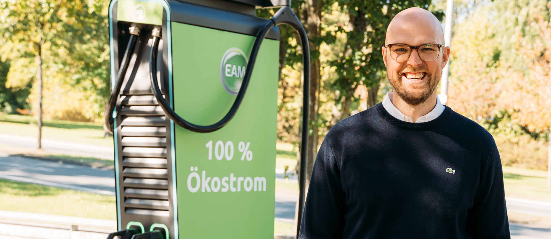 Lächelnder Mann neben einer Ladestation für Elektrofahrzeuge mit dem Aufdruck "100 % Ökostrom"" im Freien."