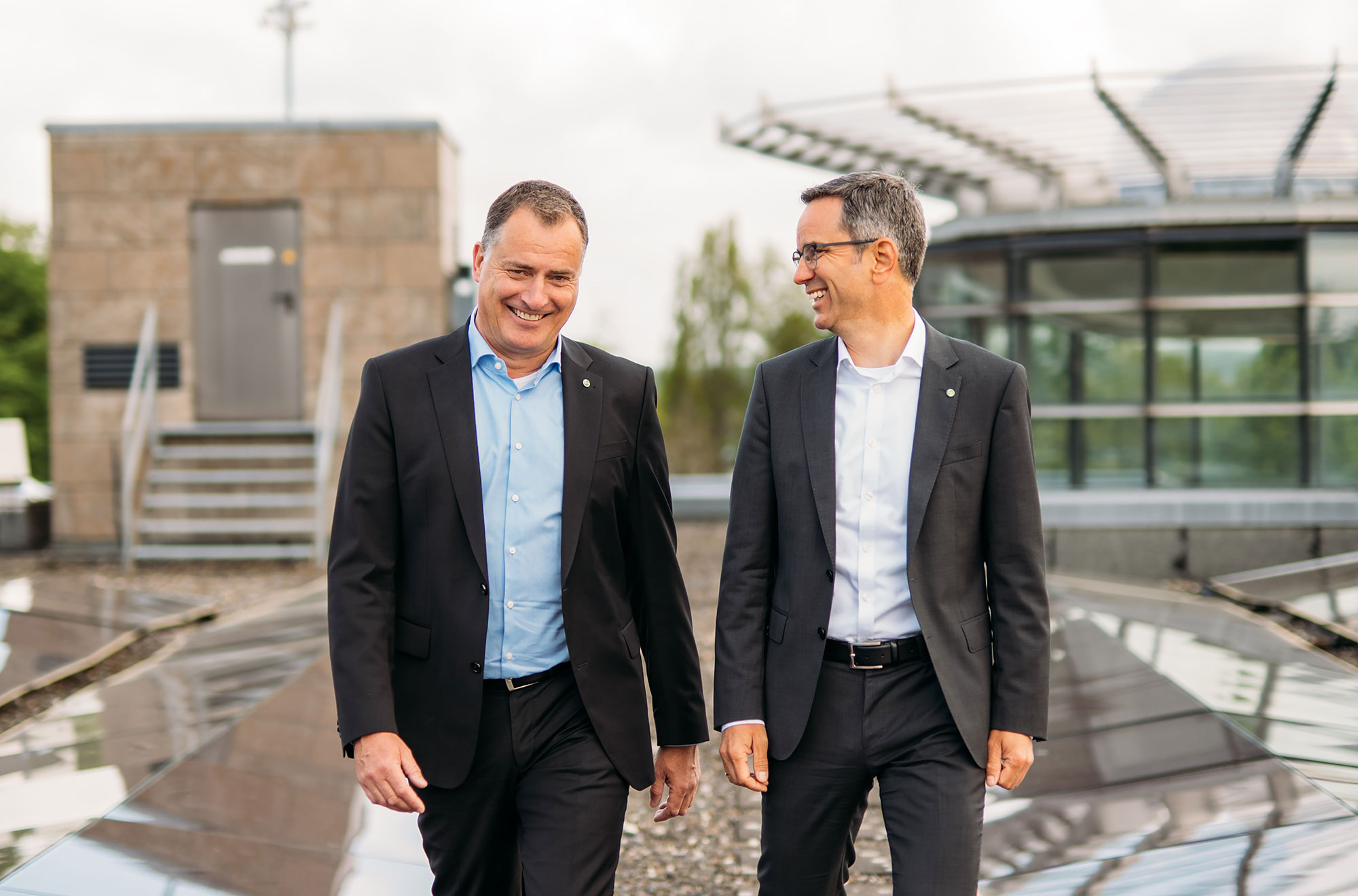Zwei Männer in Anzügen gehen lächelnd auf einem modernen, steinernen Weg mit Glasarchitektur im Hintergrund.