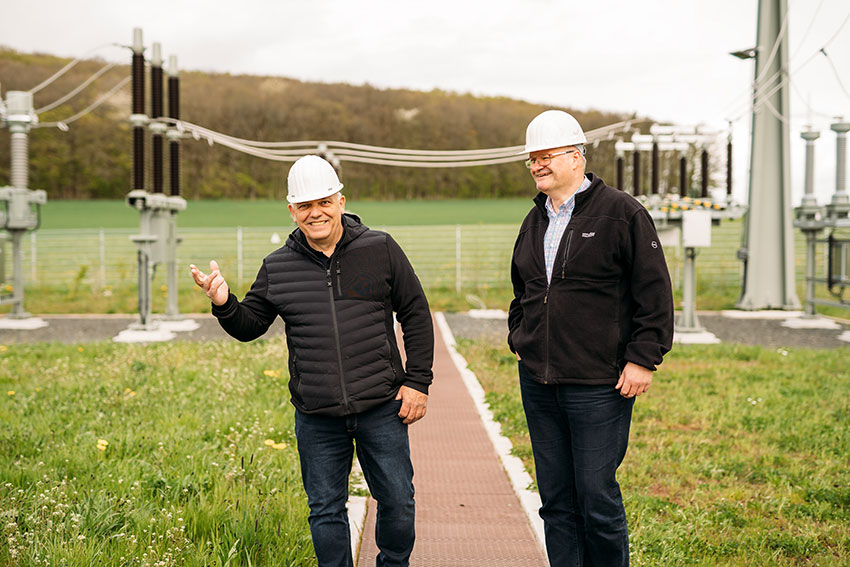 Zwei Männer in Schutzhelmen stehen auf einem Weg in einem Energieversorgungsbereich und sprechen miteinander.