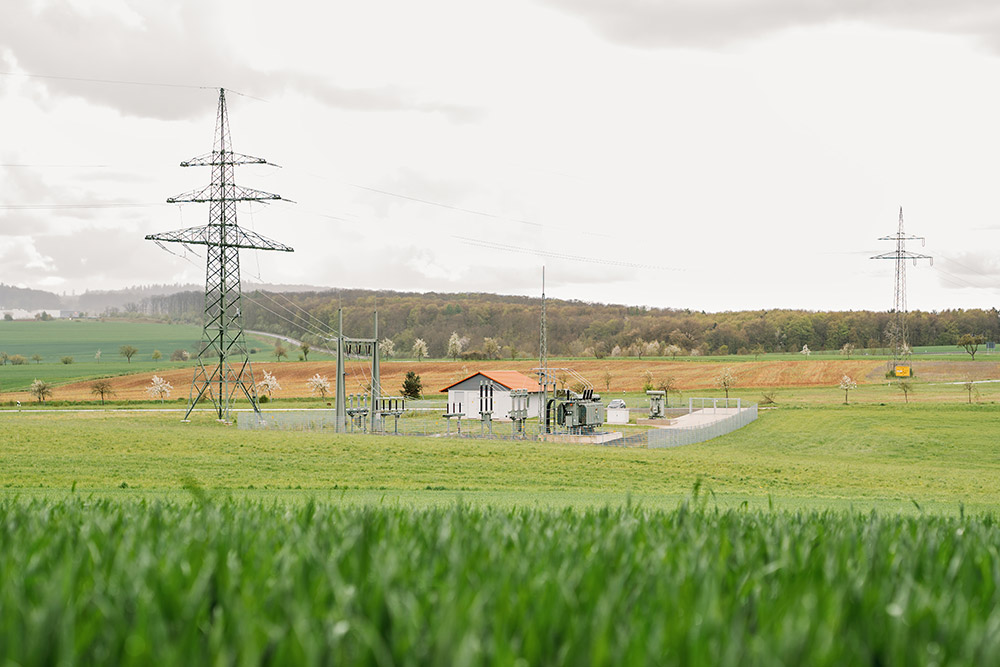 Ein Umspannwerk umgeben von Wiesen und hügeliger Landschaft, mit hohen Strommasten und einem bewölkten Himmel.