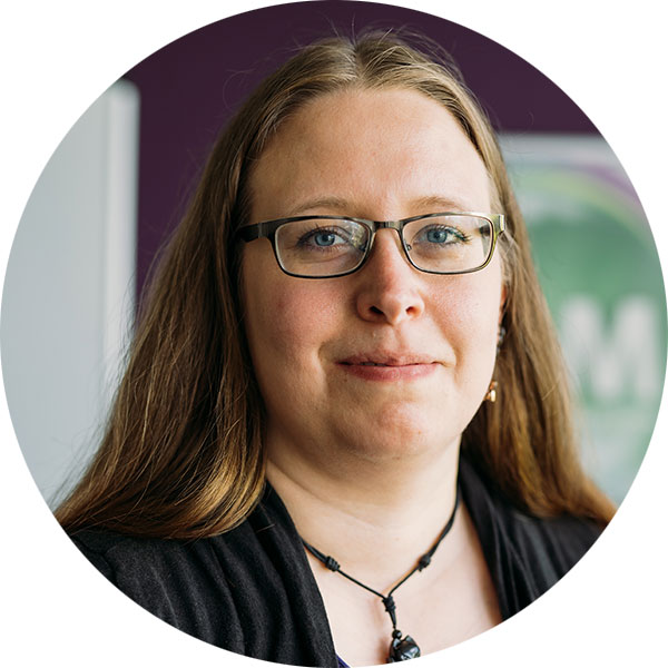 Porträt einer Frau mit langen Haaren, Brille und einem Halsband, vor einem lila Hintergrund.