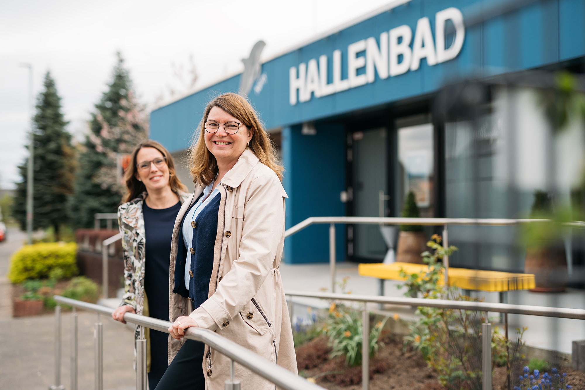 Zwei Frauen stehen an einem Handlauf vor dem Hallenbad, umgeben von Pflanzen und einer modernen Architektur.