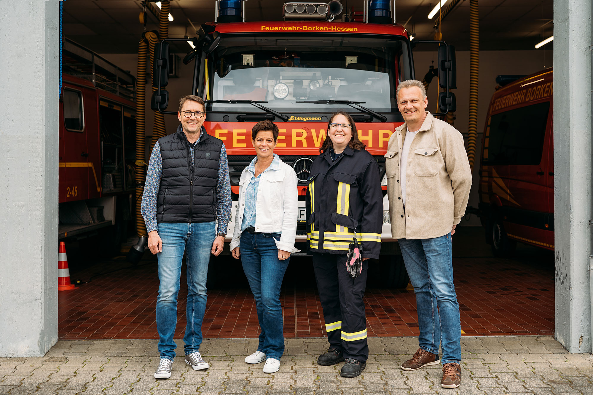 Vier Personen stehen vor einem Feuerwehrfahrzeug in einer Fahrzeughalle. Sie tragen Arbeitskleidung und lächeln in die Kamera.