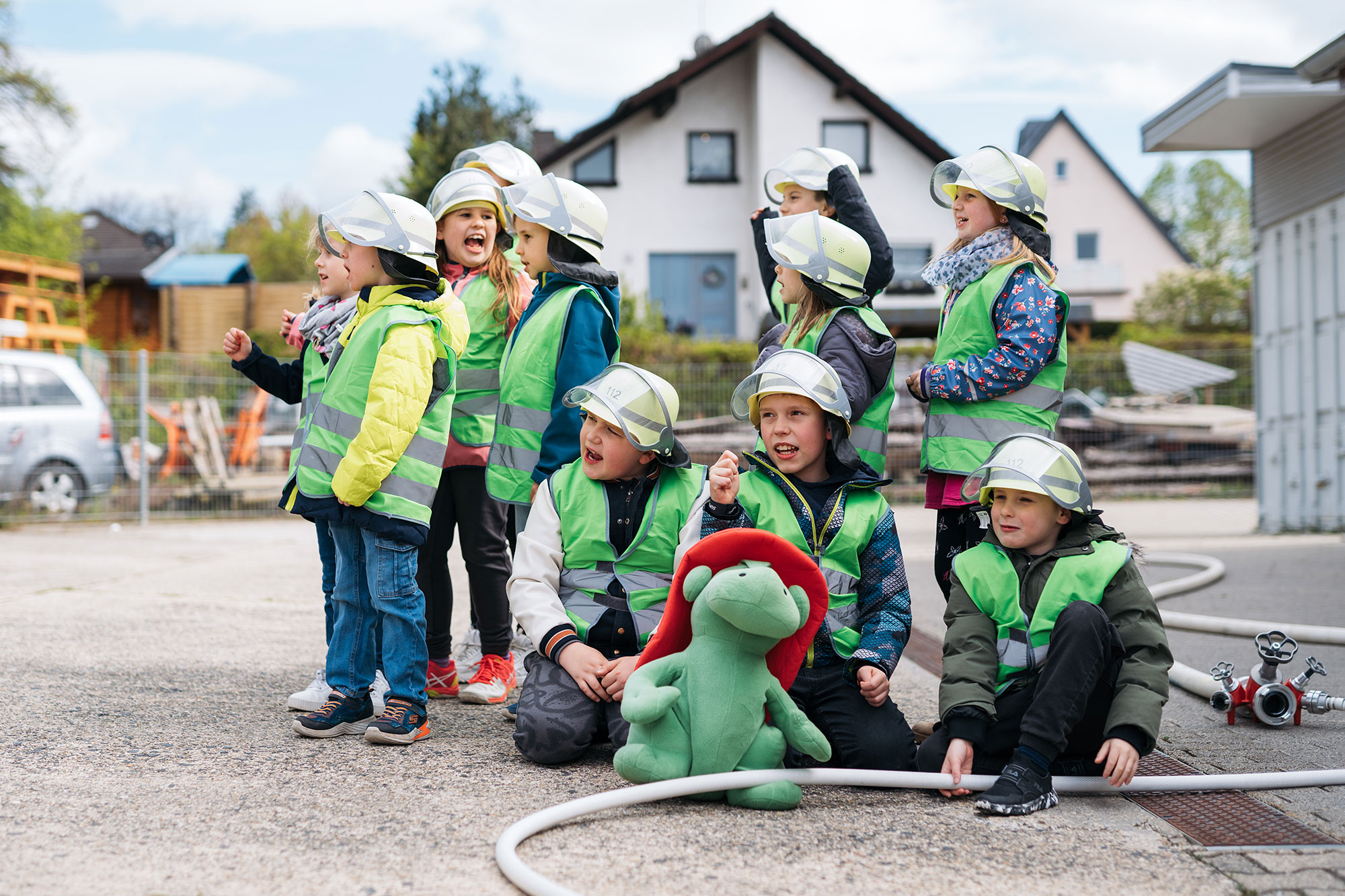 Gruppe von Kindern in Sicherheitswesten und Helmen, lachend und posierend, mit einem grünen Plüschtier auf dem Boden.