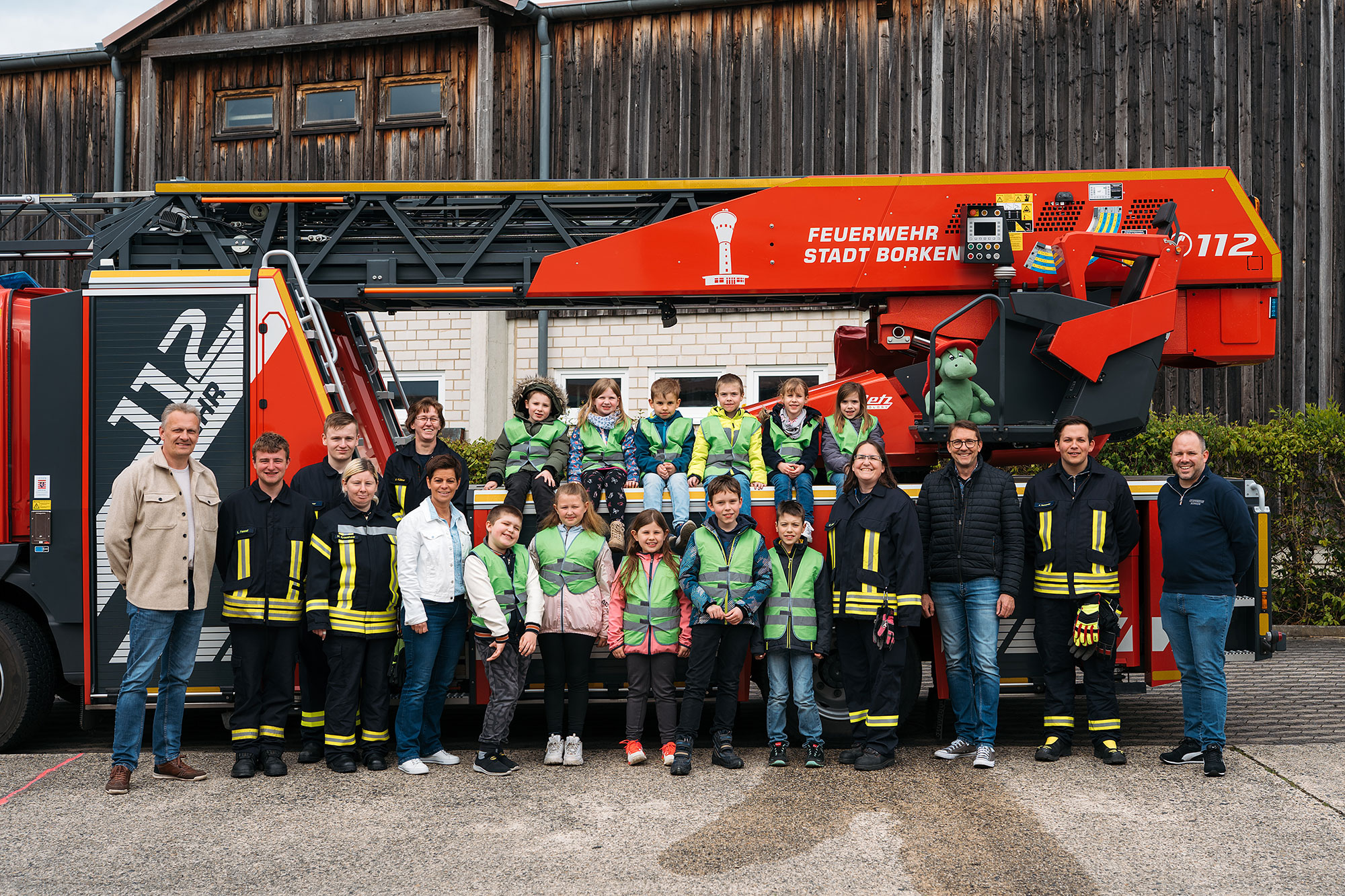 Gruppenschild mit Feuerwehrleuten und Kindern in reflektierenden Westen vor einem Feuerwehrfahrzeug.