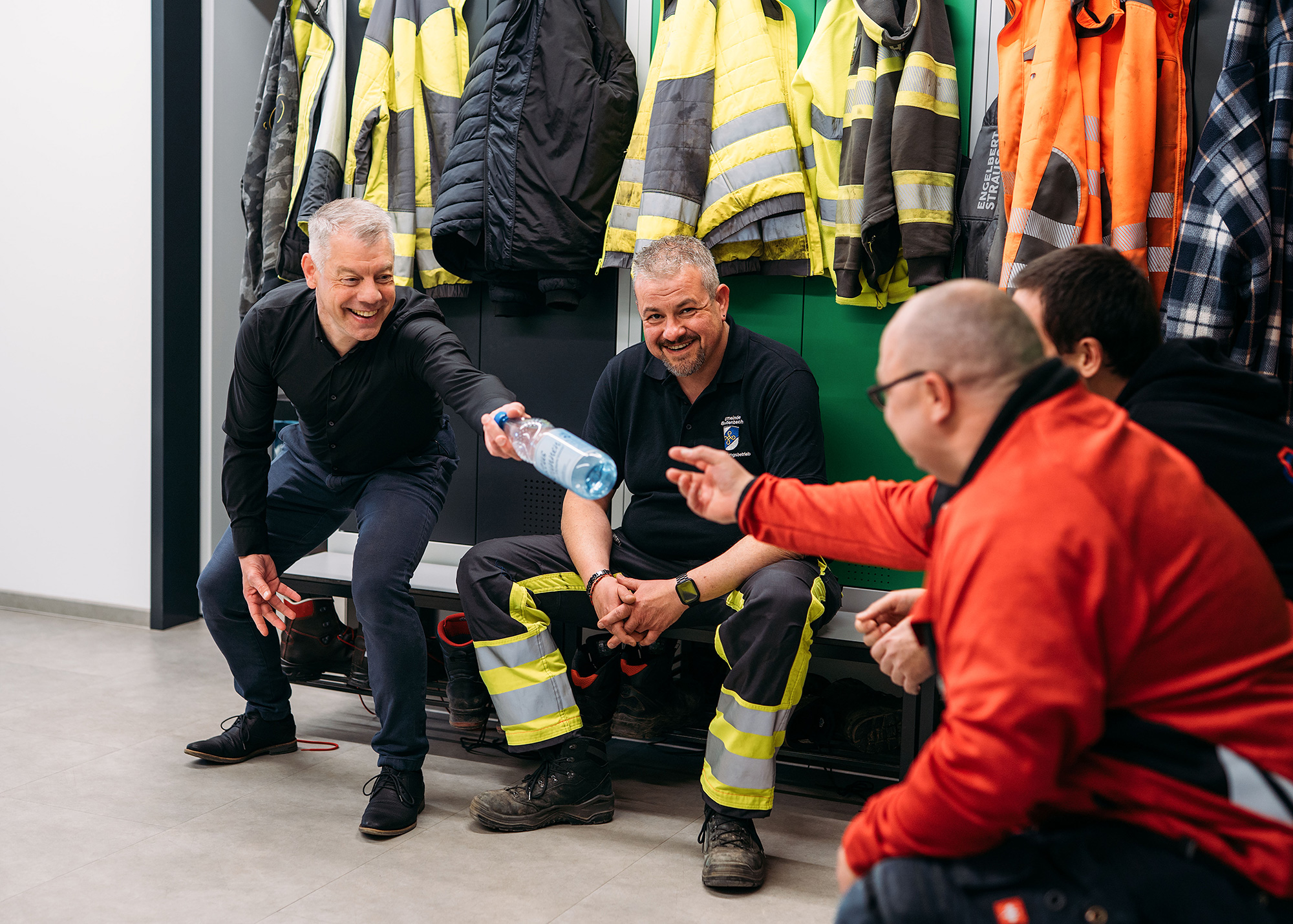 Mehrere Männer sitzen lachend in einer Umkleide vor aufgehängter Arbeits- und Warnschutzkleidung; einer reicht eine Wasserflasche weiter.
