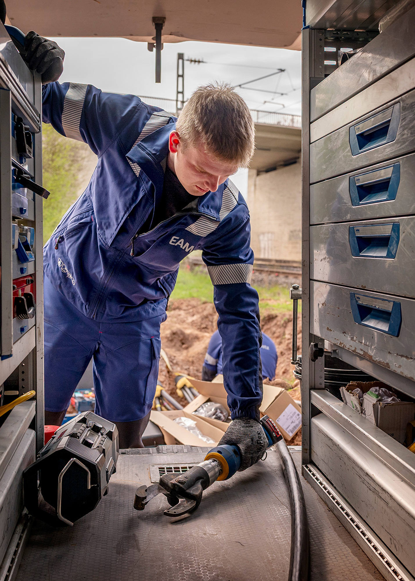 Ein EAM-Mitarbeiter in blauer Arbeitskleidung entnimmt ein Werkzeug aus einem Servicefahrzeug; im Hintergrund ist eine Baustelle mit offenem Graben zu sehen.