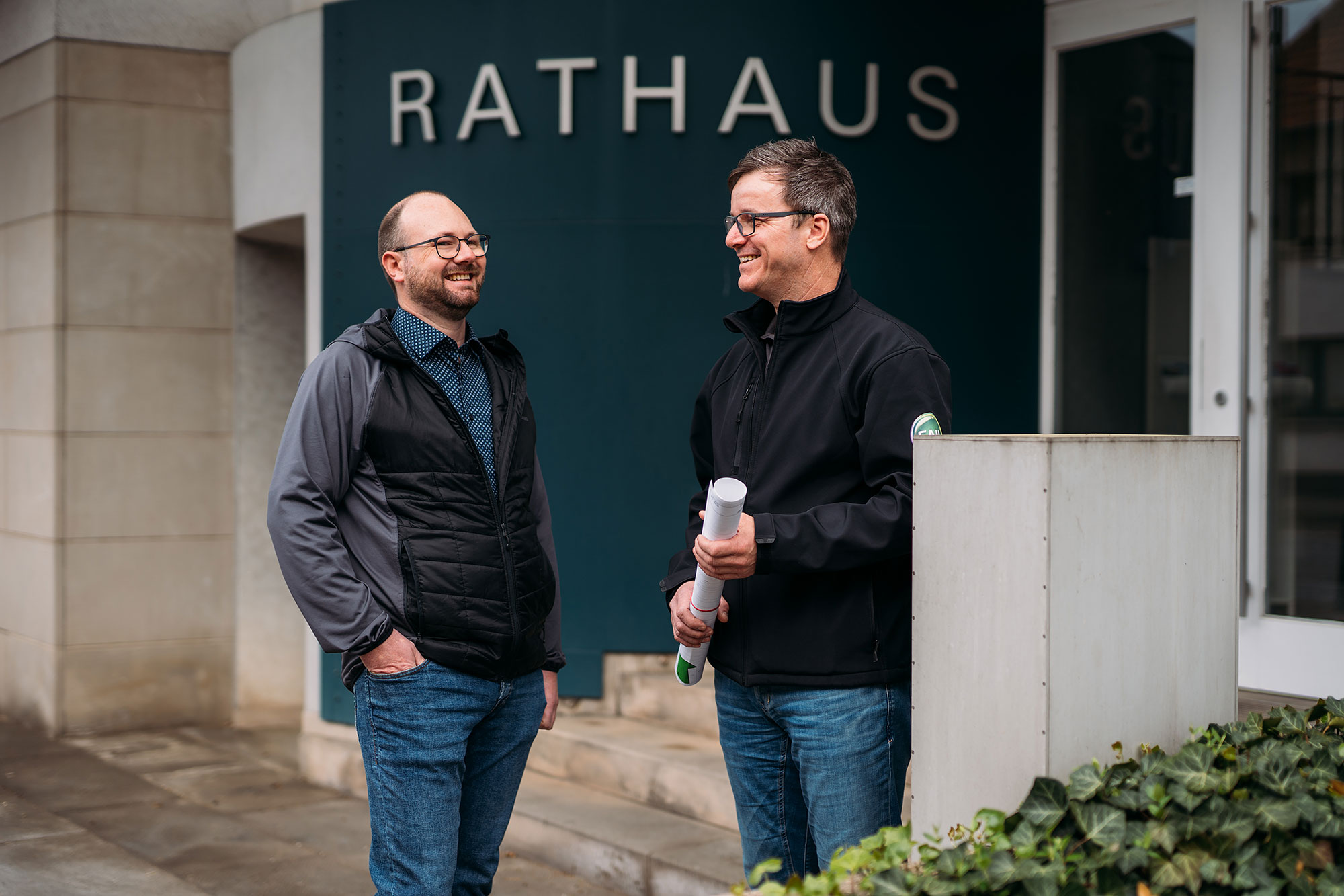 Zwei Männer stehen lachend vor dem Eingang eines Rathauses; einer hält zusammengerollte Pläne in der Hand.