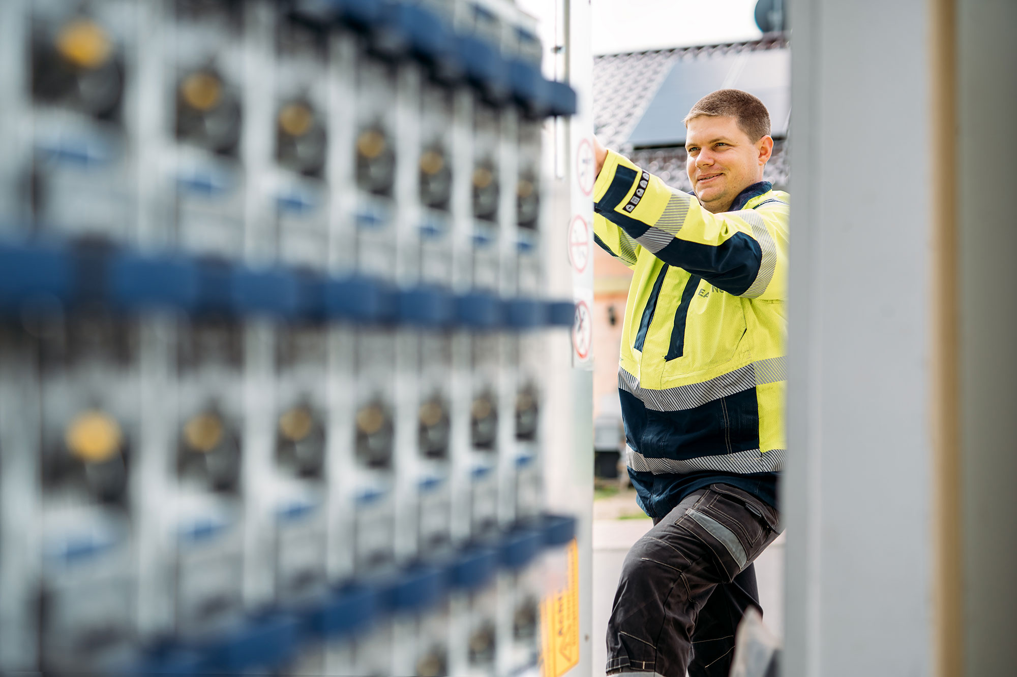 Ein EAM-Mitarbeiter in gelber Warnschutzjacke arbeitet an einer geöffneten technischen Anlage; links im Vordergrund sind unscharf Stromzähler zu sehen.