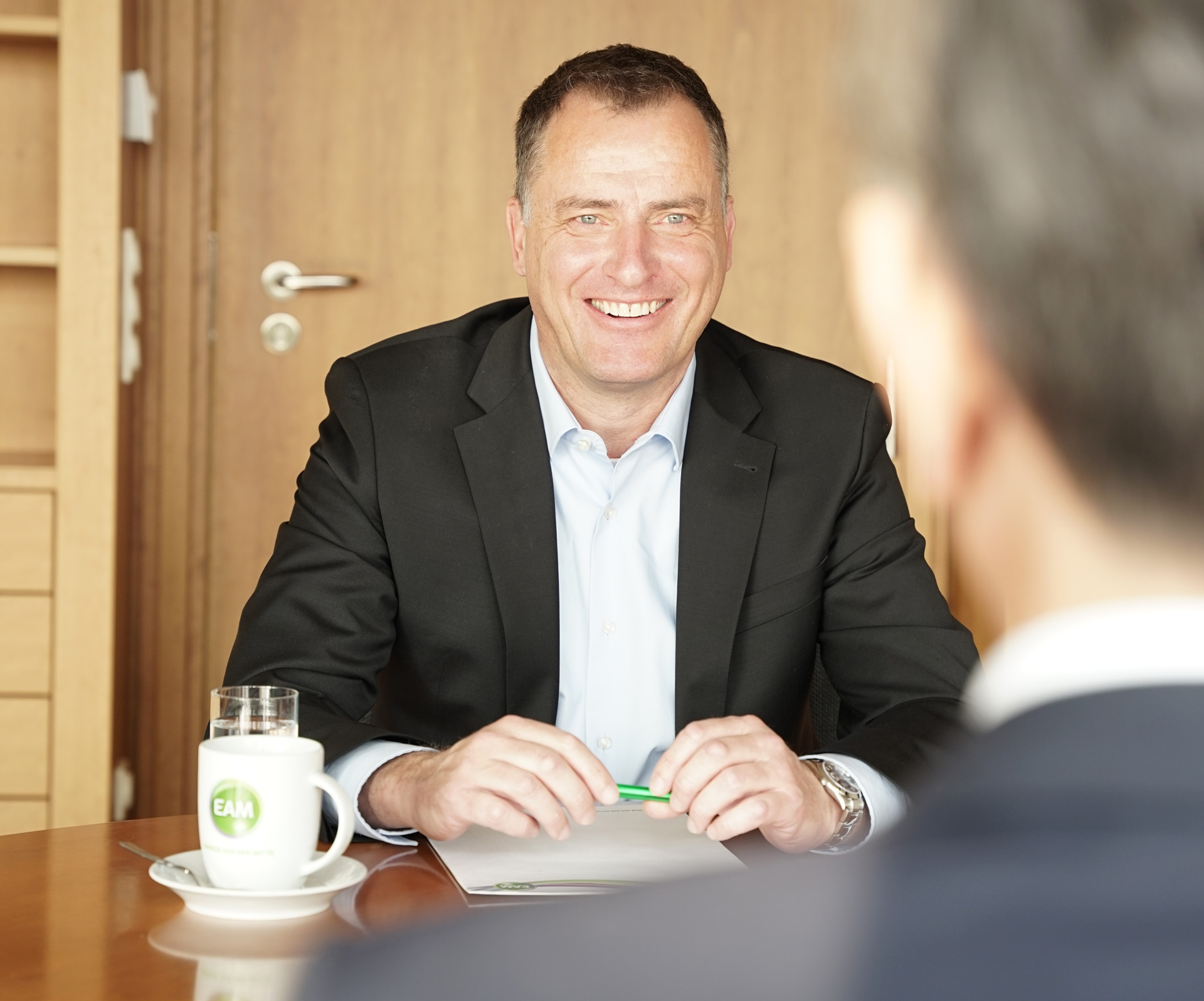 Lächelnder Mann in Anzug, sitzt am Tisch mit einer Tasse Kaffee und einem Stift, Blickrichtung zur Kamera.