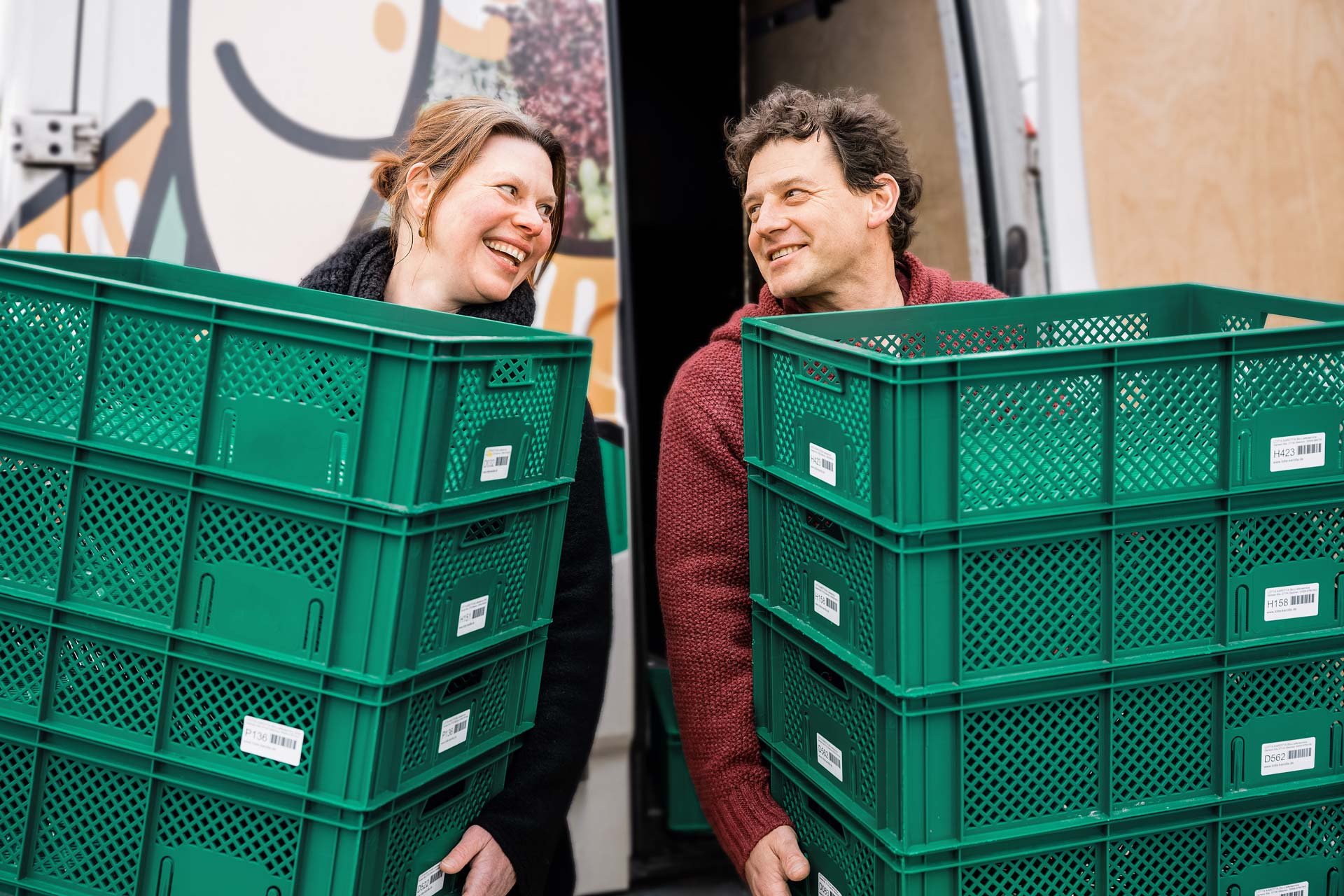 Zwei Personen lächeln einander an, während sie grüne Kisten vor einem Lieferwagen tragen.