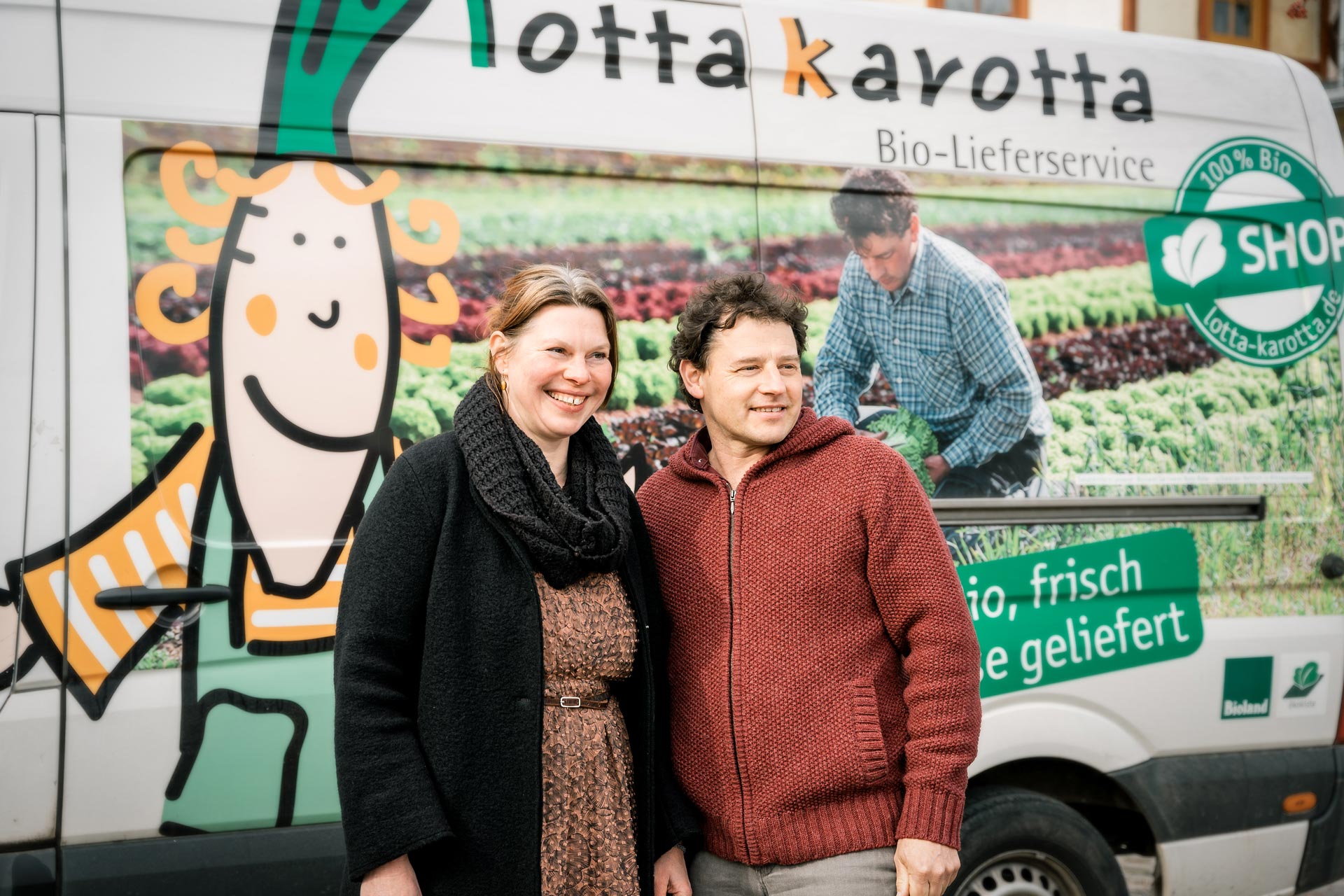 Zwei lächelnde Personen stehen vor einem Lieferwagen mit buntem Bio-Lieferdienst-Design.
