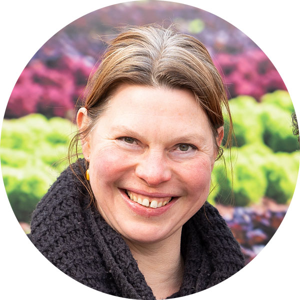 Lächelnde Frau mit langen, braunen Haaren in einem schwarzen Schal, vor einem bunten Gartenhintergrund.