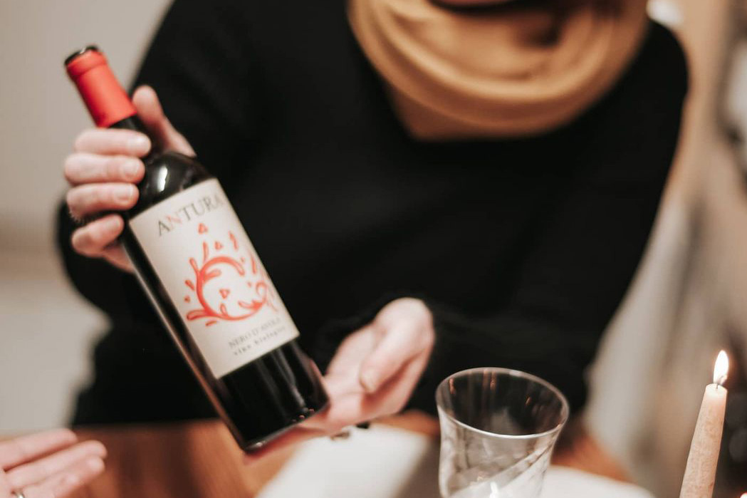 Eine Person hält eine Flasche Rotwein mit dem Etikett „Antura“ in der Hand, während eine Kerze brennt.