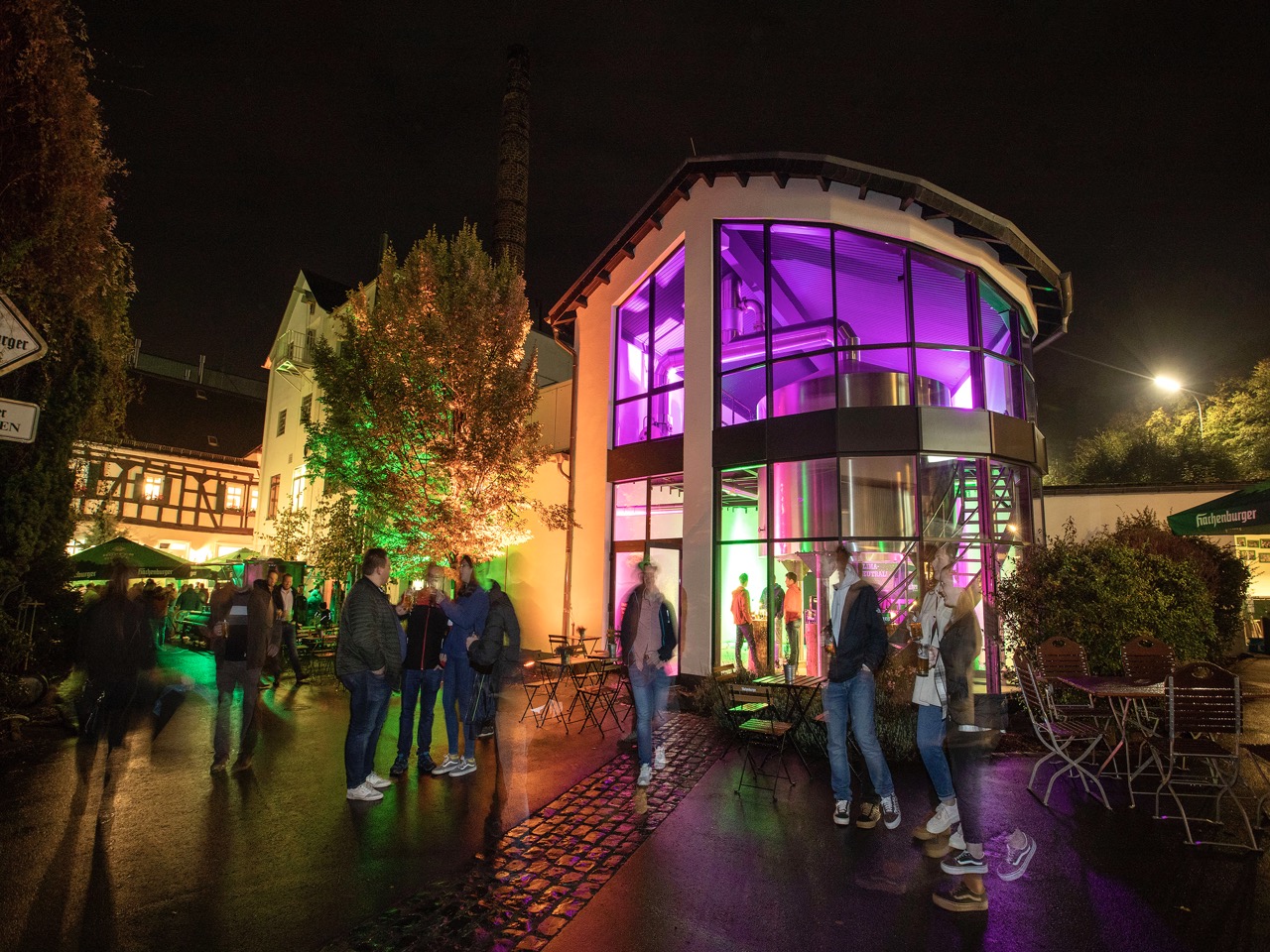Menschen stehen in der Nacht vor einem modern beleuchteten Gebäude mit Glasfassade und Sitzelementen im Freien.